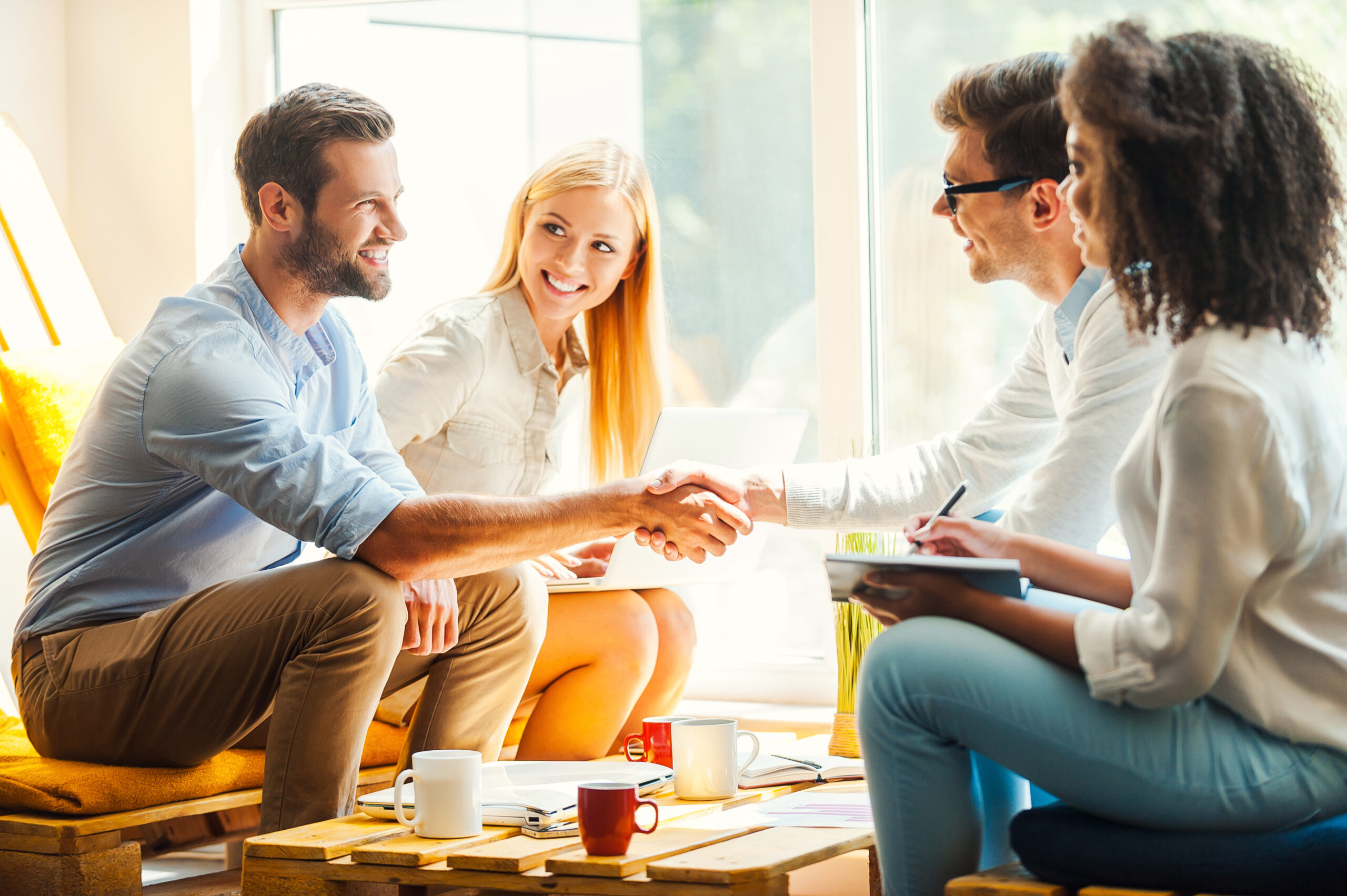 Welcome to our team! Two happy young womenworking togetherat the rest area while two men shaking hands and smiling